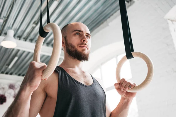 Strong bearded athlete holding crosstraining rings at cross hall. Muscular man holding gymnastic rings at light gym and looking away
