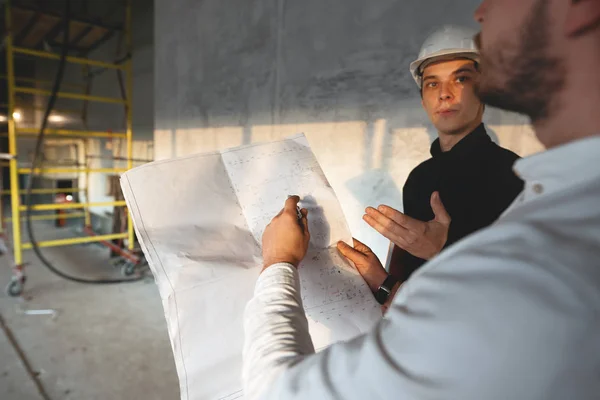 Safety inspector and build worker inside building under construction. Engineer hold blueprint in hand and discussing project with builder on construction site