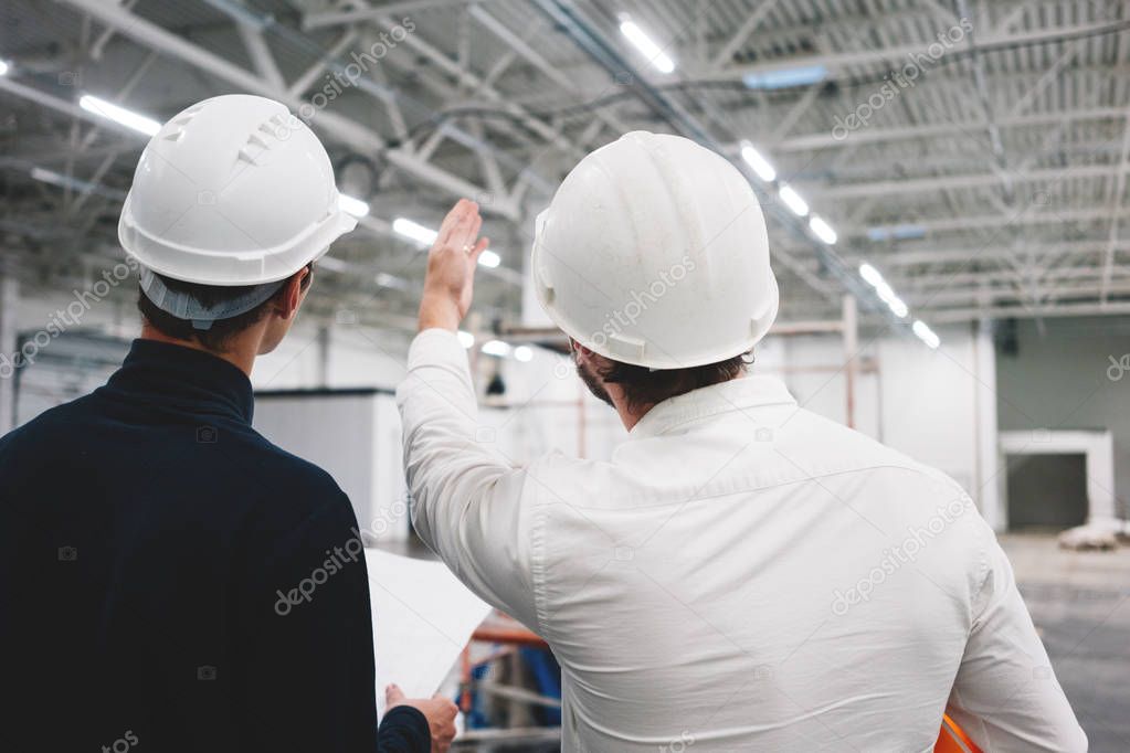 Two building engineers wearing safety hard hat discussing blueprint on construction site. Worker and inspector have meeting inside building under construction