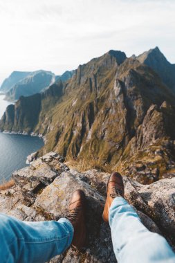 Denizin üzerindeki dağın kenarında oturan tek başına gezgin. Büyük dağlar ve yürüyüş botları üzerinde Pov görünümü.