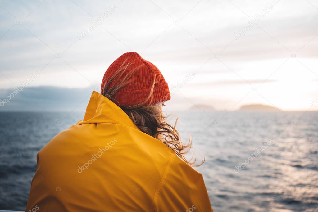 Young traveler wearing red hat and yellow raincoat floating on ship looking at sunset sea after storm and foggy mountains on skyline. Lifestyle travel, scandinavian authentic landscap