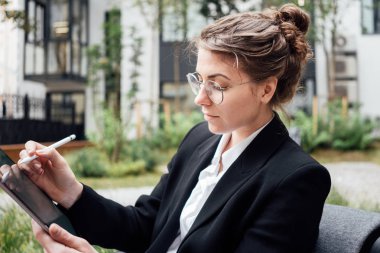 Verandada oturan kafede pensive iş kadını dijital tablet tutun ve içme suyu. Kahve dükkanında dijital tablet üzerinde çalışan gözlük takan güzel kadın