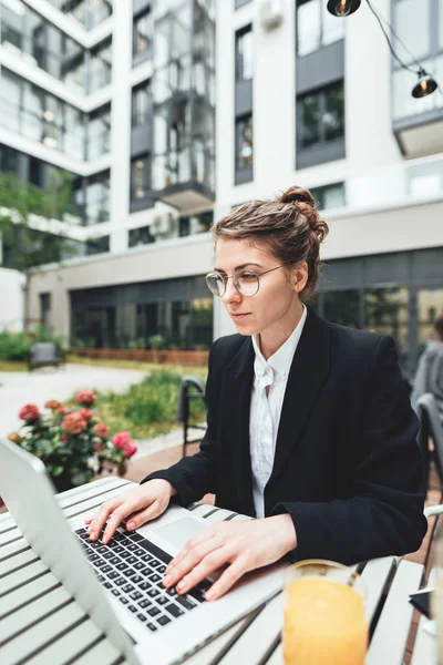 Portrait of female freelancer wearing glasses with computer. Young business woman sitting at coffee shop on veranda and working on laptop