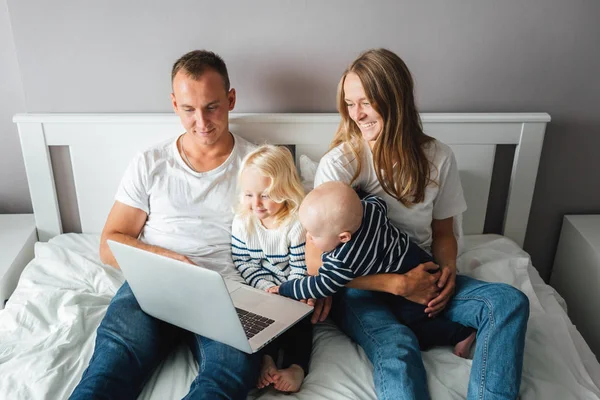 Happy family sits on bed with children using the computer together at home. Young dad and mother have fun with her son and daughter