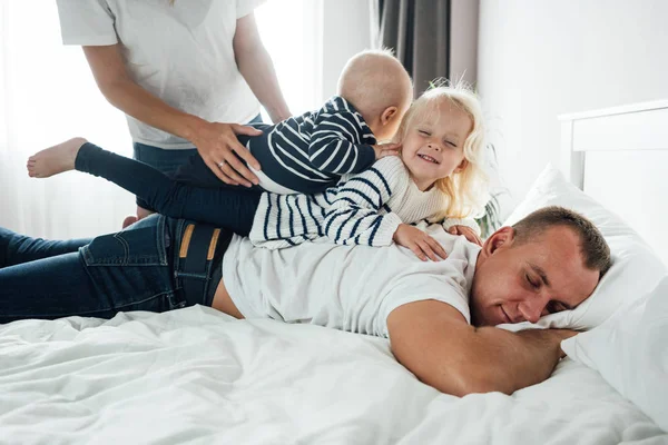 Tired father sleep but his children interferes him. Young happy father lying on bed while his daughter play with him