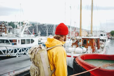 Turistik sırt çantalı, sarı ceketli, otantik balıkçı tekneleri arasında yürüyen bir turist. Sırt çantalı hippi gezgin gezgin, İskandinav şehrini keşfediyor.