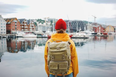 Sırt çantalı, sarı paltolu ve radikal şapkalı, İskandinav şehrine bakan kaygısız bir gezgin. Gemiler arasında küçük otantik şehirde yürüyen bir turist