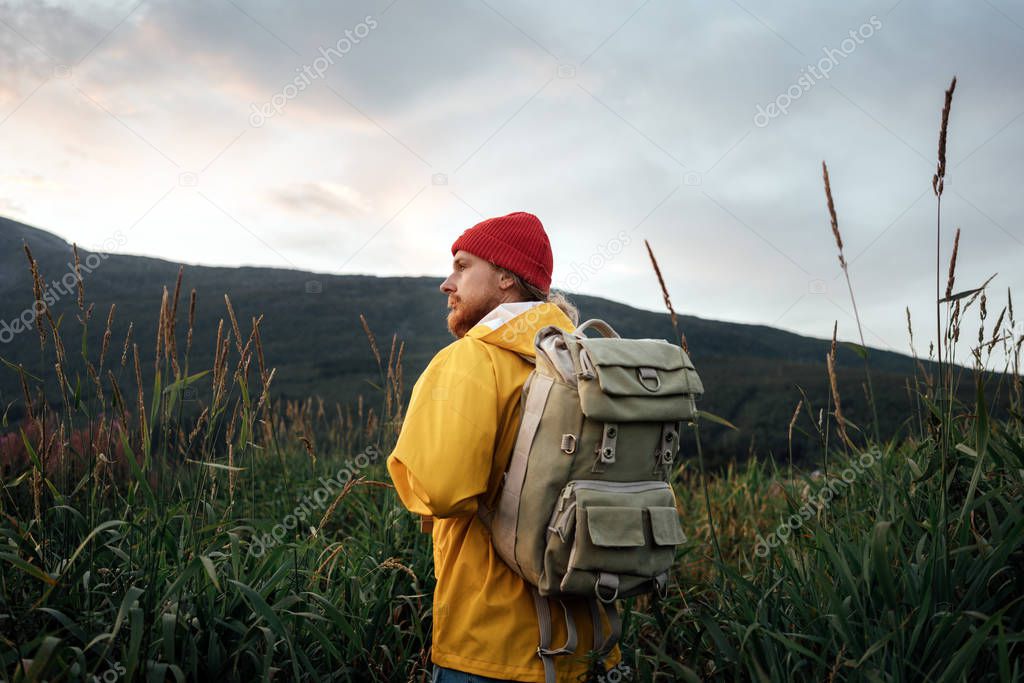 Back view of man tourist with backpack standing in front of the mountain massif while journey by scandinavian. Male traveler wearing yellow jacket explore national park and hiking outdoor landscape