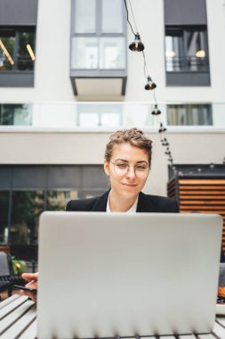 Veranda 'daki kafede oturan ve sosyal medya projesinde çalışmak için kişisel dizüstü bilgisayar kullanan yaratıcı kadın portresi. İş kadını terastaki kafede oturuyor ve bilgisayarla çalışıyor.