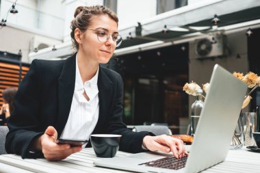 Kafe verandasındaki düşünceli iş kadını dijital sosyal medyada çalışmak için kişisel laptop kullanıyor. Akıllı telefondan mesaj yazan güzel bir bayan serbest yazar.