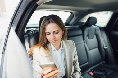 İş kadını arka koltukta arabayla seyahat ederken akıllı telefon kullanıyor. İş kadını telefona bakıyor ve yazı yazıyor.