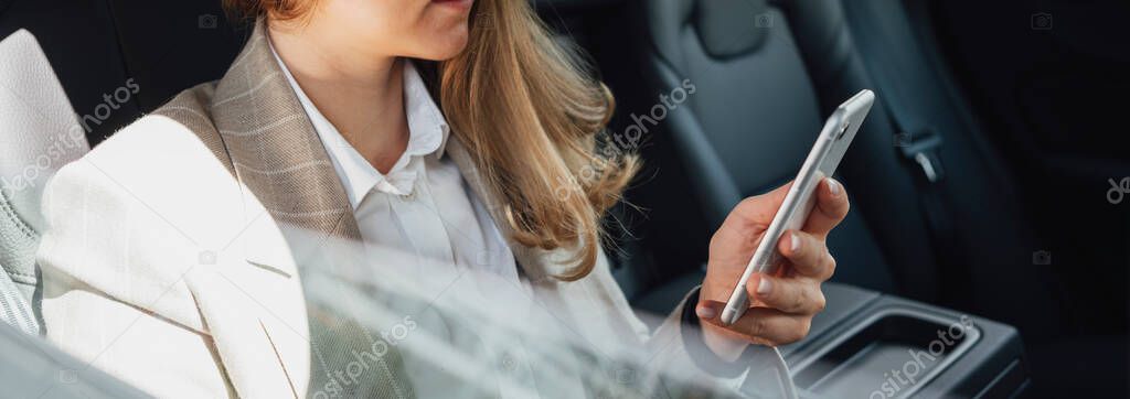 Young smarty business woman hold smartphone in hand while sits in car on backsit. Business lady using smartphone while traveling by car. Wide image
