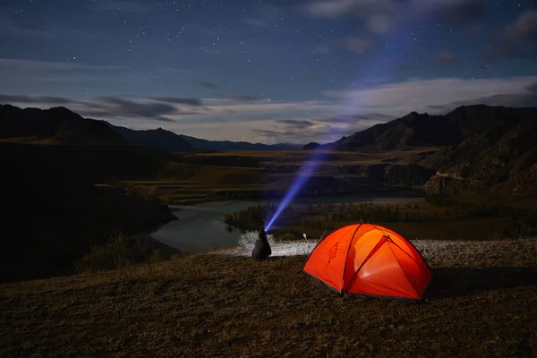 Een toerist horloges de sterrenhemel in de buurt van de tent op de achtergrond van een mooi en kronkelende rivier. — Stockfoto