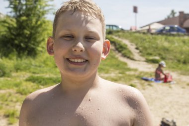 Kumsalda durup gülümseyen sarı saçlı çocuk.