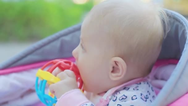 Μικρό νεογέννητο κοριτσάκι με ξανθά μαλλιά κάθεται σε ένα καροτσάκι με ένα παιχνίδι — Αρχείο Βίντεο