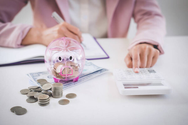 Business woman putting coins into piggy bank, writing to report and using calculator to analysis business investment strategy with income of money coin and dollar, financial concept.