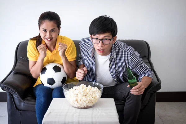 Young Asian couple love watching soccer match on tv and cheering football team, celebrating with beer and popcorn at home, sports and entertainment concept.