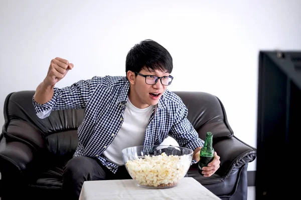 Young Asian Man fanclub watching soccer match on tv and cheering football team, celebrating with beer and popcorn at home, sports and entertainment concept.