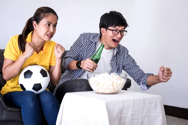 Young Asian couple love watching soccer match on tv and cheering football team, celebrating with beer and popcorn at home, sports and entertainment concept.