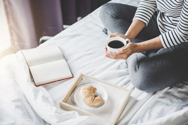放松和娱乐 年轻的幸福妇女在床上与老书和晨杯咖啡在手和牛角面包 — 图库照片