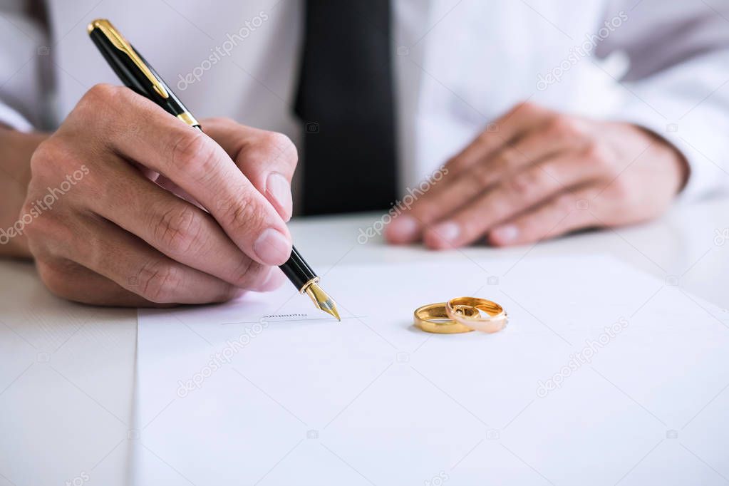 Hands of husband signing decree of divorce (dissolution or cancellation) of marriage filing divorce papers and two golden marry ring.