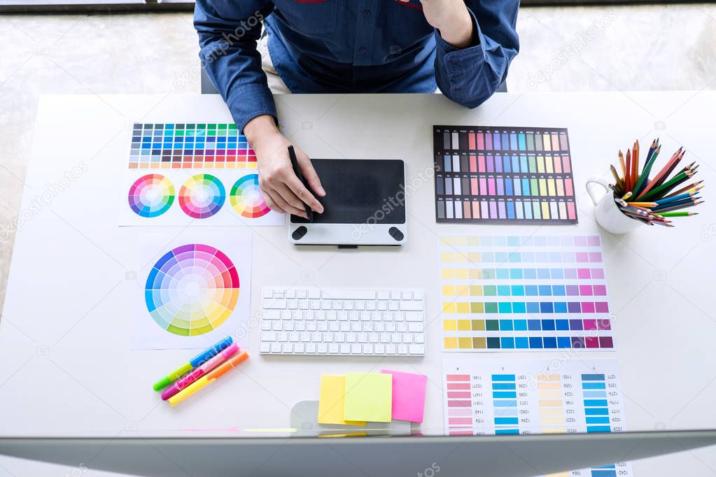 Image of male creative graphic designer working on color selection and drawing on graphics tablet at workplace with work tools and accessories, top view workspace.