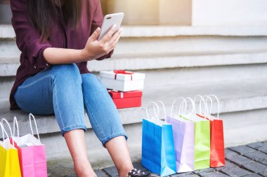 Tüketim, alışveriş, yaşam kavramı, smartphone alışveriş zevk oynarken alışveriş torbaları ve hediye kutusu yakın oturan genç kadın.