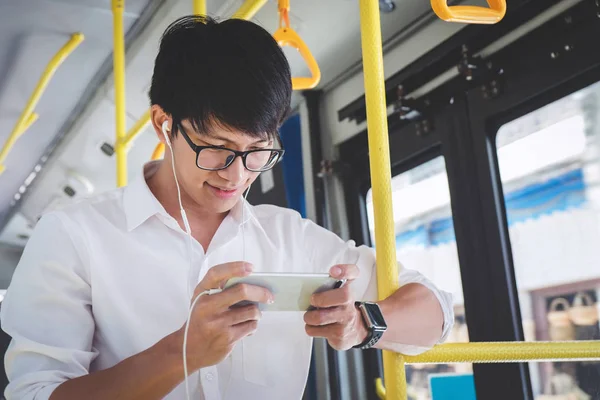 Young Asian man traveler standing on a bus using smartphone watch video or playing game while smile of happy, transport, tourism and road trip concept.