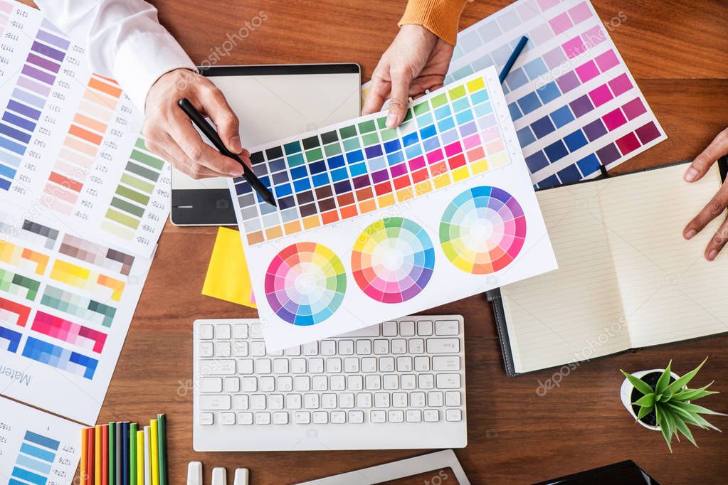 Two colleague creative graphic designer working on color selection and color swatches, drawing on graphics tablet at workplace with work tools and accessories, top view workspace.