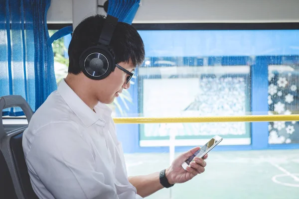 Young Asian man traveler sitting on a bus using smartphone watch video or listening music while smile of happy, transport, tourism and road trip concept.
