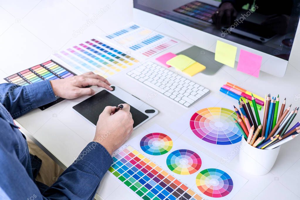Male creative graphic designer working on color selection and color swatches, drawing on graphics tablet at workplace with work tools and accessories.