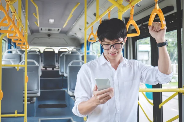Young Asian man traveler standing on a bus listening to music on smartphone while smile of happy, transport, tourism and road trip concept.