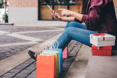 Tüketim, alışveriş, yaşam kavramı, smartphone alışveriş zevk oynarken alışveriş torbaları ve hediye kutusu yakın oturan genç kadın.