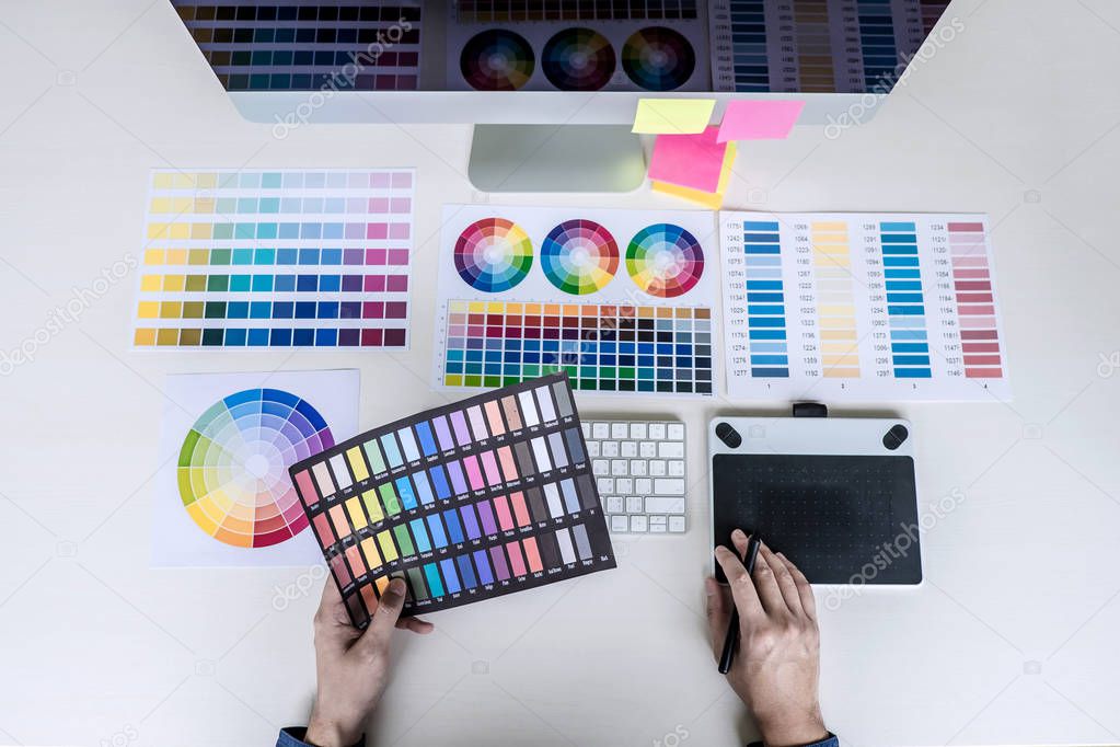 Male creative graphic designer working on color selection and color swatches, drawing on graphics tablet at workplace with work tools and accessories, top view workspace.