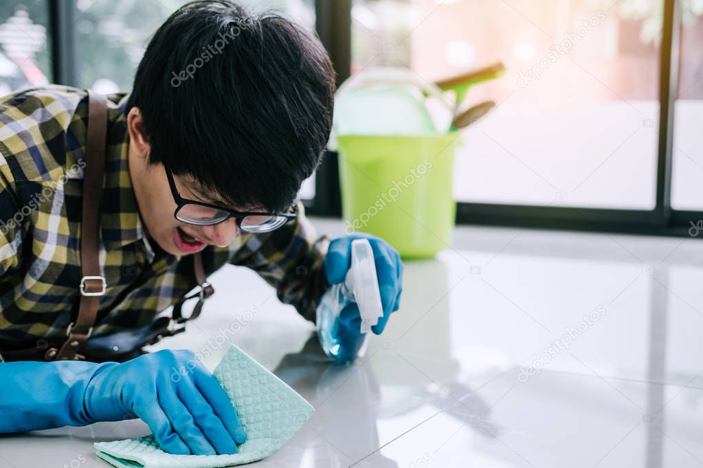 Husband housekeeping and cleaning concept, Happy young man in blue rubber gloves wiping dust using a spray and a duster while cleaning on floor at home.