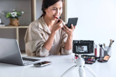 Mutlu gülümseyen zarif kadın blogcu, makyaj malzemesi kozmetik inceleme ürünü sunuyor ve kamera ekranında sosyal ağa canlı yayın yapan video bloglarını öğretiyor..