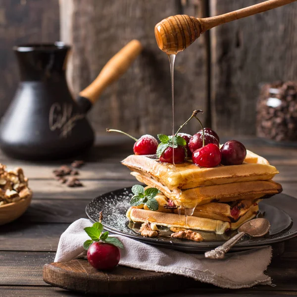 Freshly made belgian waffles with honey flows and powdered sugar. Cherries on top of waffles on wooden desk and napkin on wooden background. Coffee beans in glass jar. Turkish coffee pot for breakfast