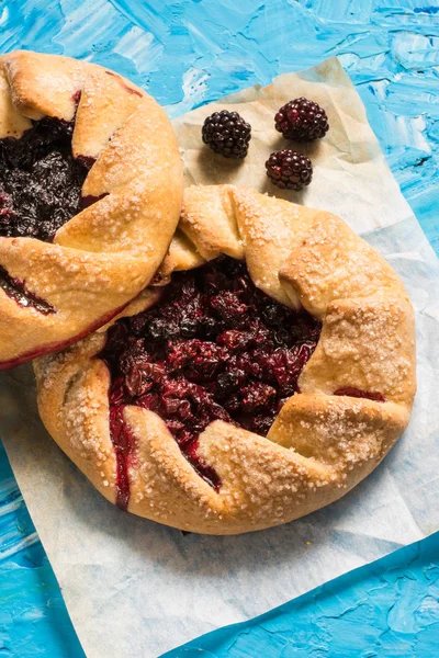 Summer galette with berries, raspberry, blueberry, blackberry, apple and crusty sugar vegetarian dough on blue background. Summer food, summer pie on parchment paper. Open pie. Vegan food.