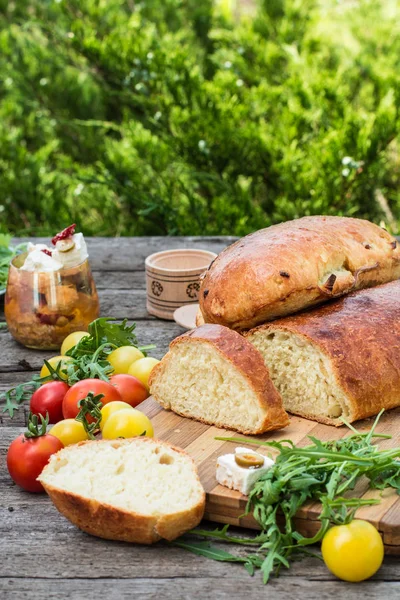 Fresh bread on wooden ground. Yellow tomato and red tomatoes with arugula. Feta cheese with olives and sundried tomatoes. Picnic, dinner outdoor. Still life of food. — Stock Photo, Image
