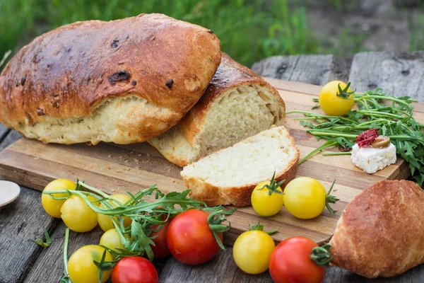 Fresh bread on wooden ground. Yellow tomato and red tomatoes with arugula. Feta cheese with olives and sundried tomatoes. Picnic, dinner outdoor. Still life of food. — Stock Photo, Image