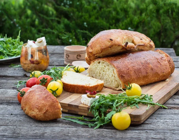 Fresh bread on wooden ground. Yellow tomato and red tomatoes with arugula. Feta cheese with olives and sundried tomatoes. Picnic, dinner outdoor. Still life of food. — Stock Photo, Image