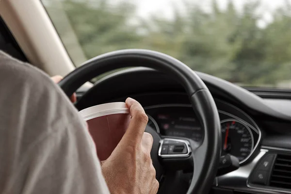 The car driver holds a coffee cup in hand, with a background of a blurred wheel