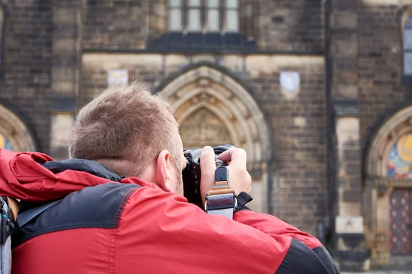 Pria berjaket merah dan dengan ransel di belakang memegang kamera dan mengambil foto bangunan tua — Stok Foto