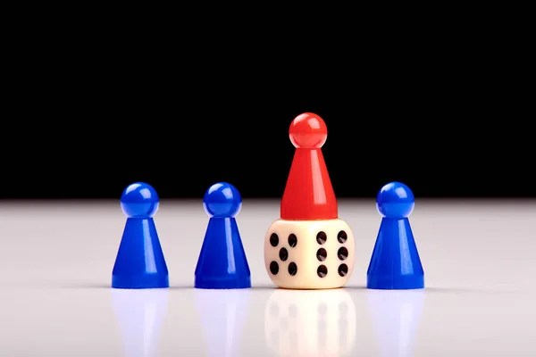 Three blue game pieces and between them one red piece stands on a dice as a winner or leader. Blurred black and white background