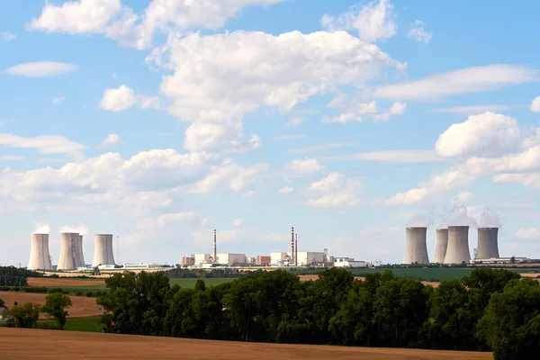 View of the smoking chimneys of a nuclear power plant in the landscape with trees, fields and houses under blue sky with clouds.