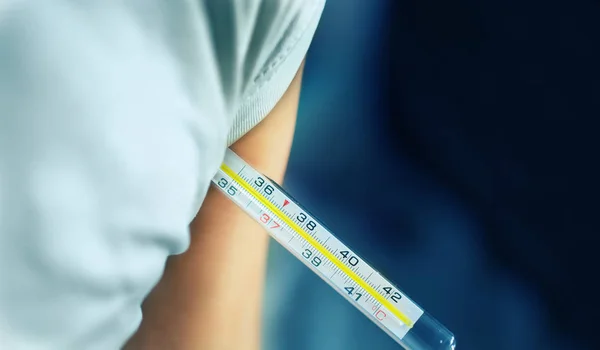 A person in white t-shirt keeps thermometer under his left arm to know body temperature, close-up with copy space on blue blurred background