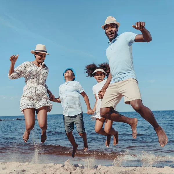 幸せな若いアフリカ系アメリカ人の家族手を繋いでいると晴れた日のビーチでジャンプ ストック写真