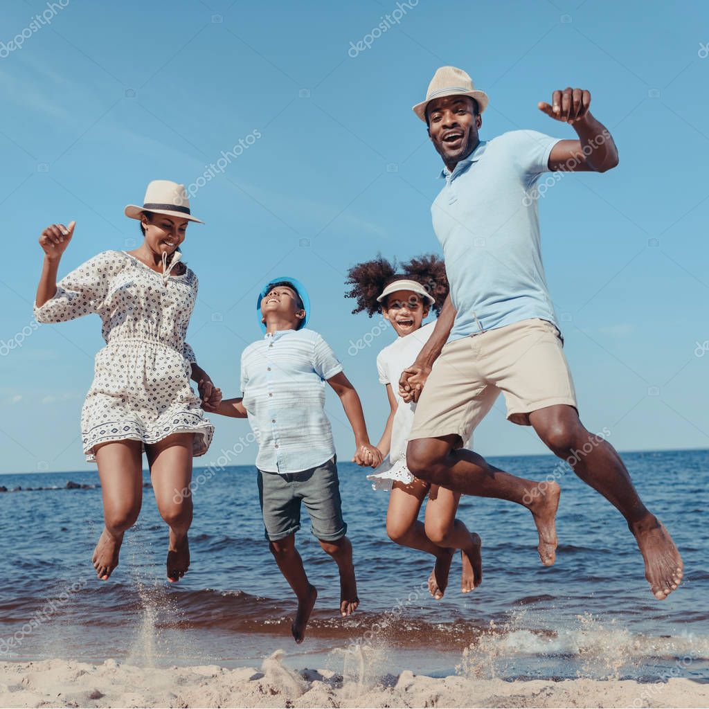Young happy african american family holding hands and jumping on beach on sunny day