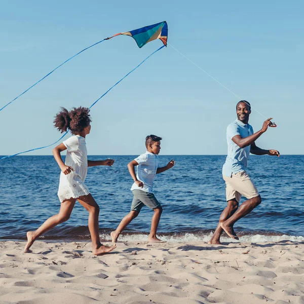 Vista lateral de feliz afroamericano padre y niños jugando con cometa en la playa - foto de stock