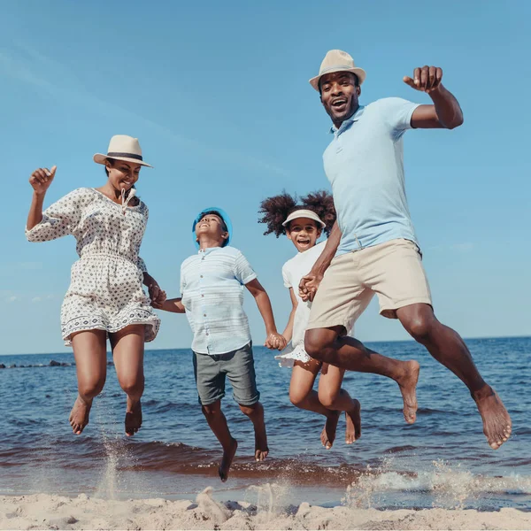 Joven feliz afroamericano familia agarrada de la mano y saltando en la playa en día soleado - foto de stock